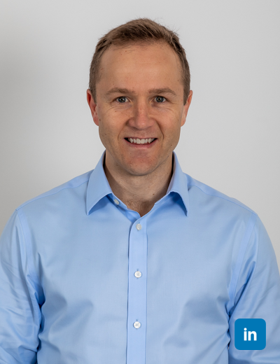 A profile image of a smiling, sandy brown haired white man wearing a light blue buttoned shirt.