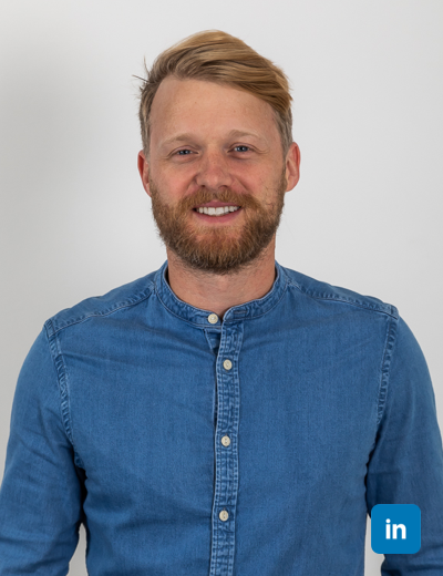 A profile image of a white man smiling with his blonde hair swept in a side parting. He wears a collarless denim buttoned shirt.