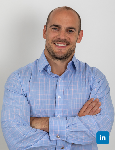 A profile image of a smiling white man with shaved head and light brown beard. He wears a light blue and pink check shirt and has his arms folded.