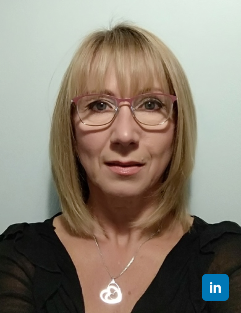 A profile image of a blonde woman with wire rimmed glasses. Her hair is neatly bobbed at her shoulders and she wears a black top with a silver pendant necklace.