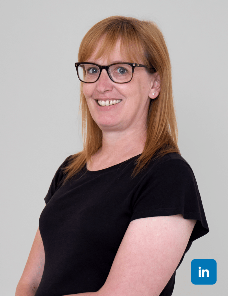 A profile image of a smiling white woman with shoulder length red hair and fringe. She wears black rimmed glasses and a black short sleeved top.