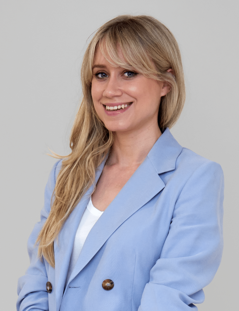 A profile image of a smiling white woman with long light blonde hair over her right shoulder and fringe. She has smokey eye make up and wears a pale blue blazer over a white top.