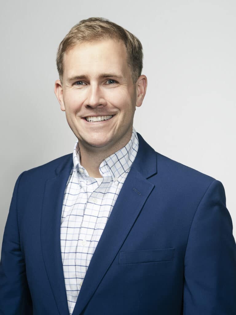 A profile image of a smiling blonde white man with a checked white and blue shirt with a navy blue suit jacket.