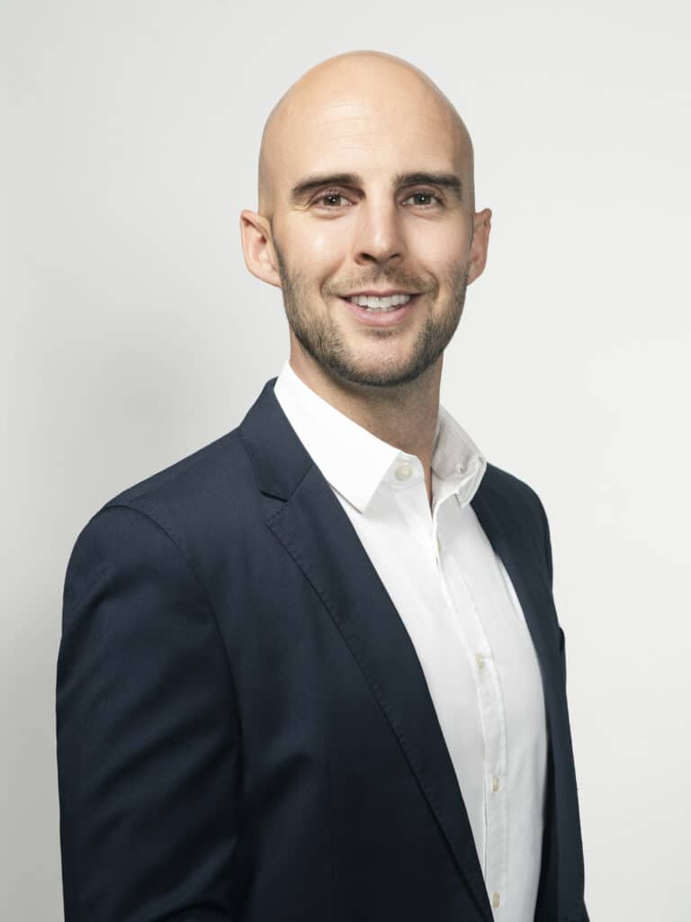 A profile photo of a young white man with shaved head and light beard wearing a white buttoned shirt and navy suit jacket.